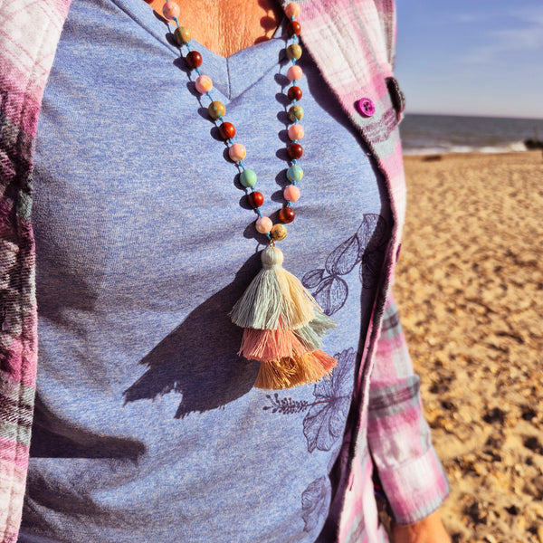 A person standing on a sandy beach wearing a colorful beaded necklace with a tassel pendant over a heathered blue-gray t-shirt and a pink plaid shirt.