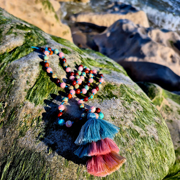A colorful beaded necklace with tiered blue, pink, and orange tassels, draped over a mossy green rock with a rocky shoreline background.