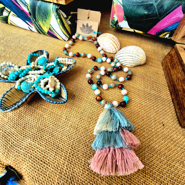 A handmade beaded necklace with turquoise, white, and brown beads, featuring a large layered tassel in light blue, teal, and pink. The necklace is displayed on a burlap surface next to seashells and a starfish-shaped decorative piece.