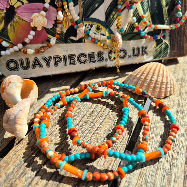 Colorful beaded bracelets with turquoise, orange, and white beads arranged on a rustic wooden surface with seashells, with a branded display card in the background reading QUAYPIECES.CO.