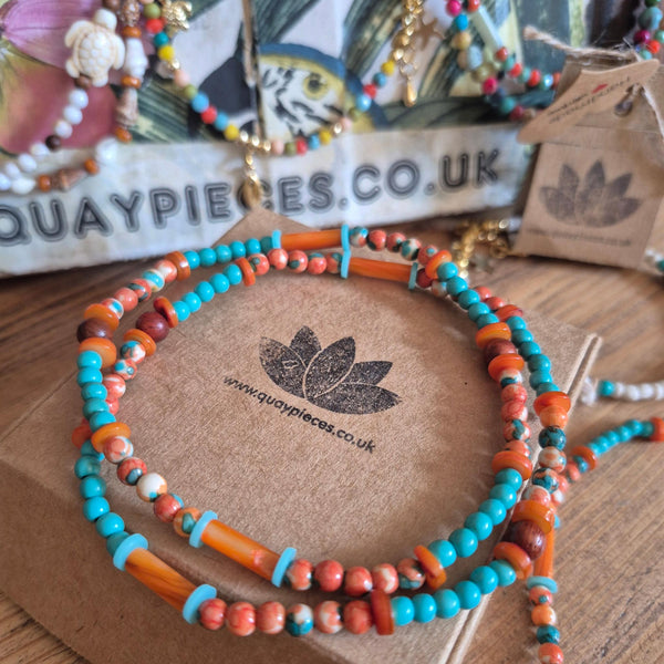 A beaded wrap bracelet with turquoise blue, orange, and coral beads displayed on a brown cardboard box. The brand name 'Quaypieces' is visible on the box and in the background.