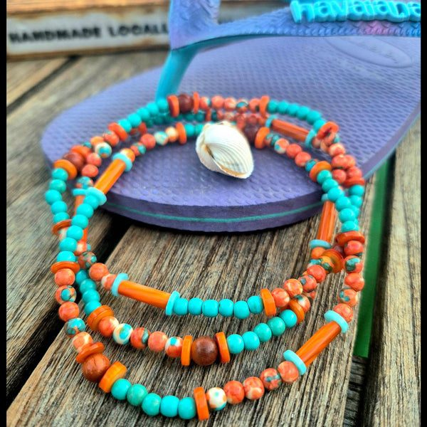 A multi-strand beaded necklace with turquoise, orange, and brown beads, featuring a small white seashell charm, displayed on a purple flip-flop sandal over a rustic wooden surface.