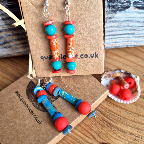 Colorful beaded drop earrings displayed on brown kraft gift boxes on a wooden surface, with a small shell dish holding red beads nearby.