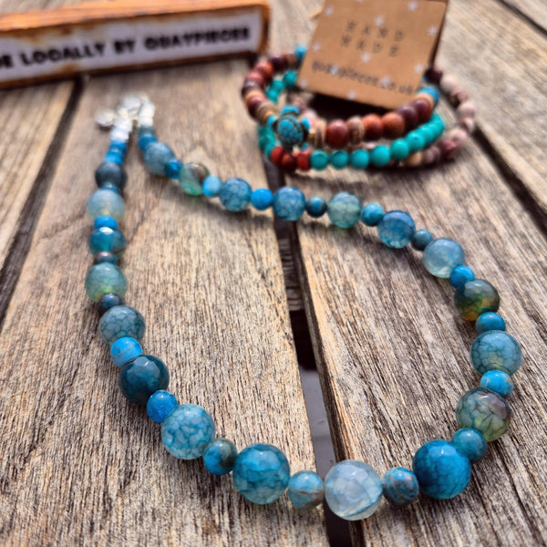 A beaded necklace made of marbled blue and turquoise beads is displayed on a rustic wooden surface, with matching bracelets in the background. A small sign in the corner reads 'Locally by quaypieces' and a card on the bracelets reads 'Hand Made'.