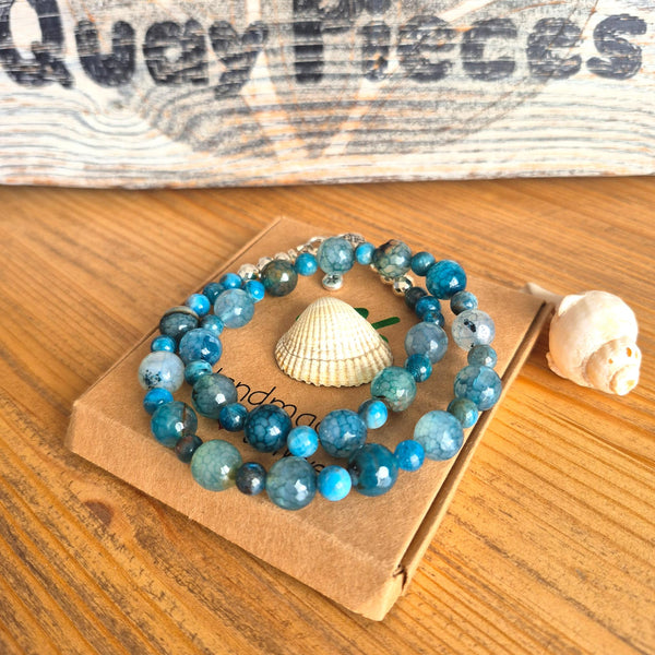 A beaded strand bracelet made of faceted blue, teal, and turquoise stones, displayed on a small kraft paper card with a seashell on a wooden surface.