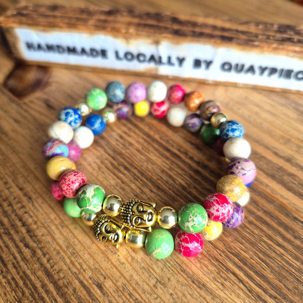 A colorful beaded bracelet with marbled beads in green, pink, blue, yellow, purple, and white, accented by two gold Buddha head charms and small metallic spacer beads, displayed on a wooden surface.