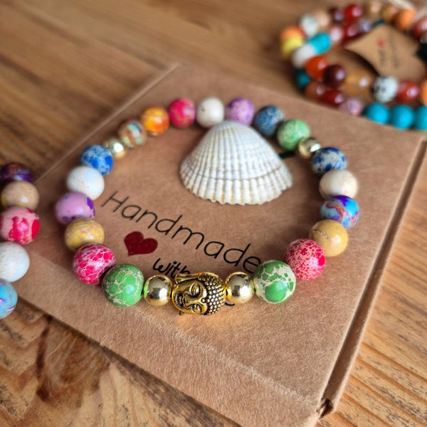 A handmade beaded bracelet with colorful marbled and solid beads, a gold Buddha charm, and small gold spacer beads, displayed on a brown kraft paper card with a seashell on a wooden table.