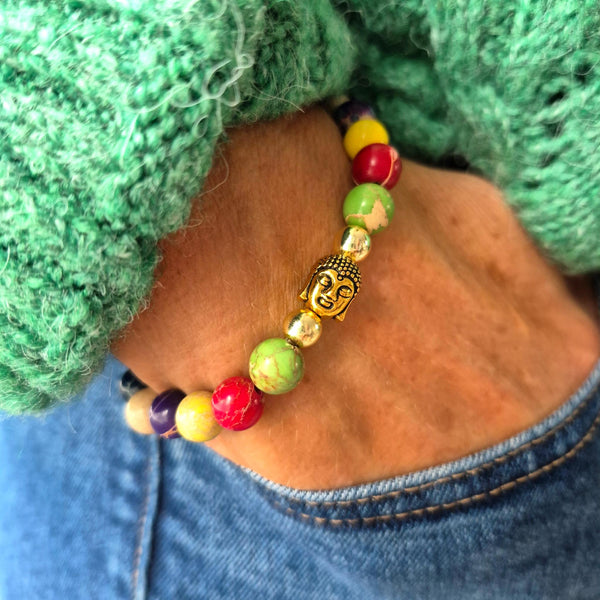 A close-up of a beaded bracelet worn on a wrist, featuring a central gold Buddha head bead surrounded by multicolored round beads in red, yellow, green, and purple, styled with a green knit sweater and blue jeans.