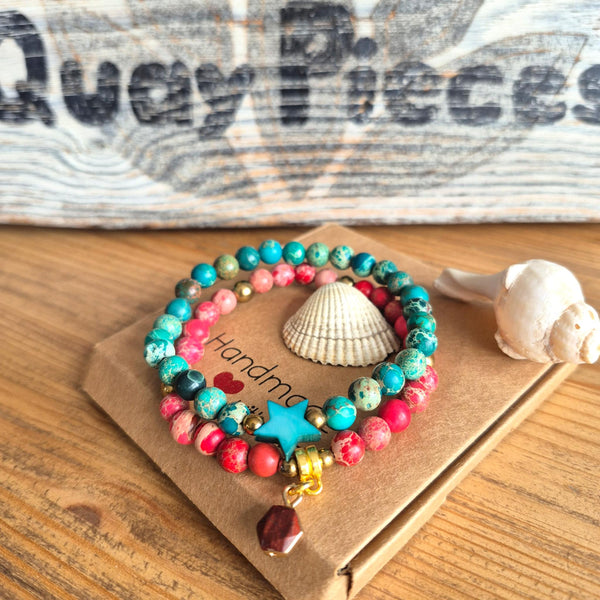 A set of colorful beaded stretch bracelets with turquoise, pink, red, and green beads, a blue star charm, and a gold bell, displayed on a brown kraft paper card with seashells on a wooden table.