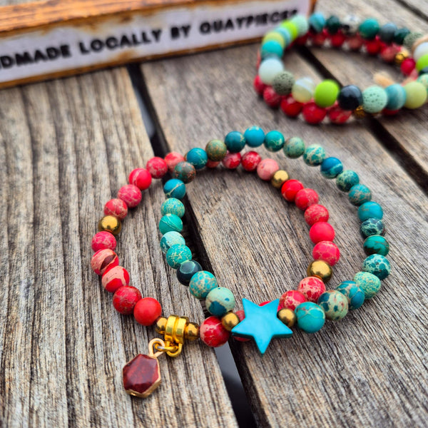 Two colorful beaded stretch bracelets displayed on a rustic wooden surface. The front bracelet features red, turquoise, and green beads with a light blue star charm and gold spacer beads, while the back bracelet is partially visible in the background.