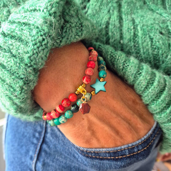 A close-up of a person's wrist wearing two beaded bracelets. One bracelet features red and turquoise beads with a small gold charm, and the other features turquoise, dark green, and red marbled beads with a light blue star charm.
