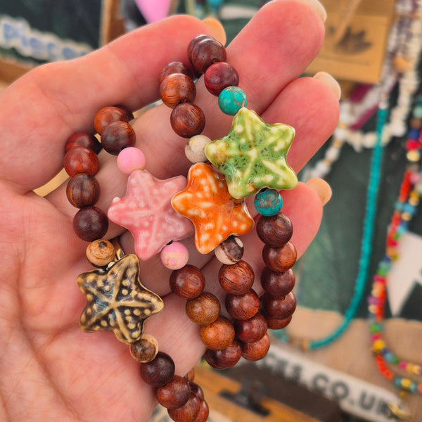A hand holding three colorful beaded bracelets with starfish charms. The bracelets feature dark brown wooden beads, small turquoise beads, a pink starfish charm, an orange starfish charm, and a green starfish charm, with one bracelet also featuring a metallic gold starfish charm.