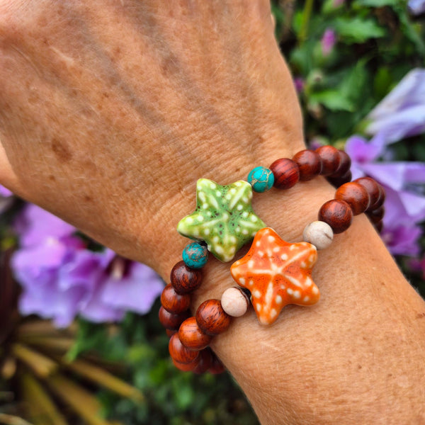 A close-up of a person's wrist wearing two beaded bracelets with starfish charms. One bracelet has dark brown wooden beads and turquoise beads, while the other has dark brown wooden beads, white beads, and an orange starfish charm.
