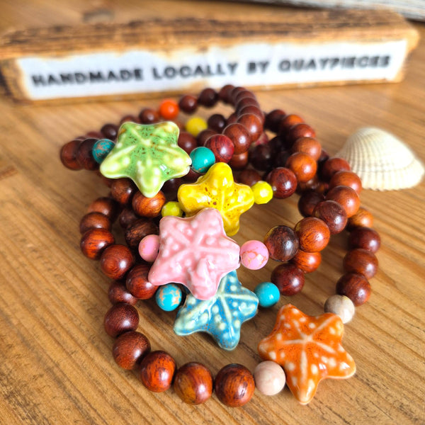 A set of handmade wooden bead bracelets with colorful starfish charms, displayed on a wooden surface with a seashell and a wooden sign in the background.