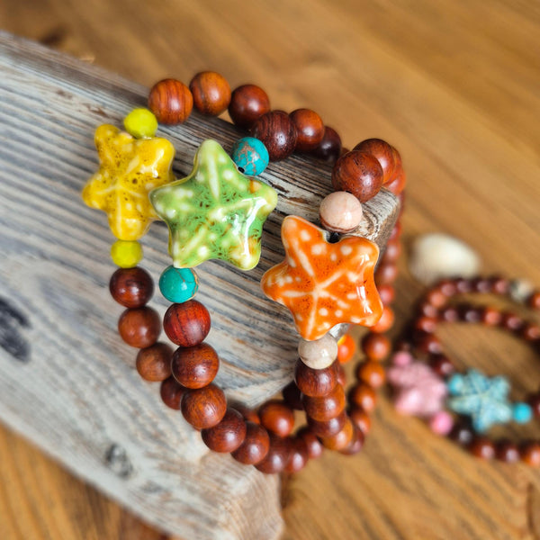 A close-up of beaded bracelets with glossy star-shaped charms in yellow, green, and orange, made with reddish-brown wooden beads, resting on a piece of weathered wood over a wooden table.