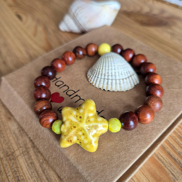 A wooden bead bracelet with a yellow starfish centerpiece and small lime-green beads, displayed on a brown kraft paper card with a seashell prop on a wooden table.