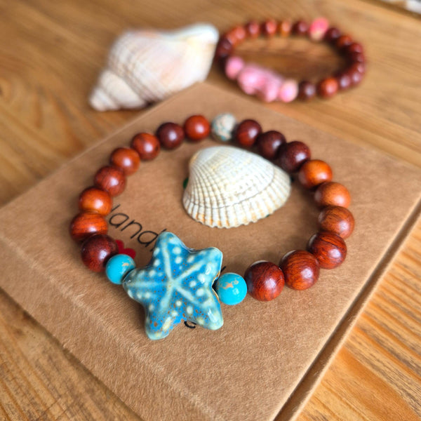 A beaded bracelet with reddish-brown wooden beads, turquoise blue spacer beads, and a blue starfish charm, displayed on a brown kraft paper card with seashells on a wooden table.