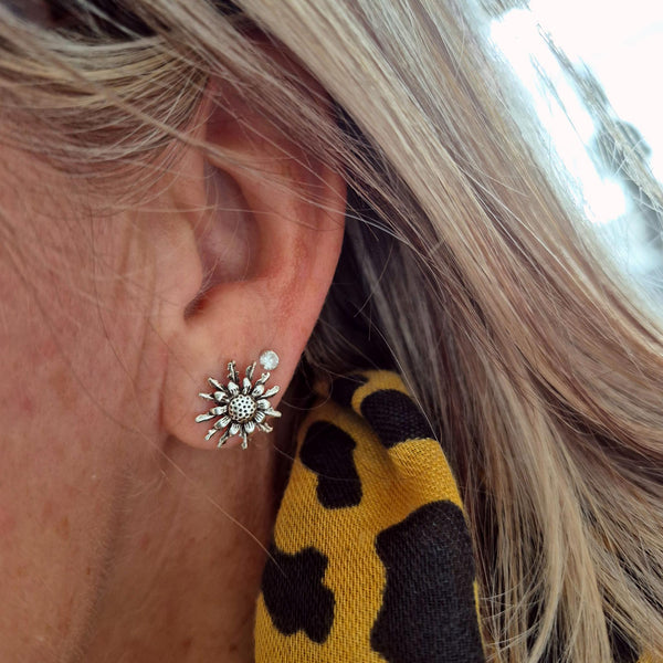 A close-up of a person's ear wearing a silver sunburst stud earring with small clear stones, next to a yellow and black patterned scarf.