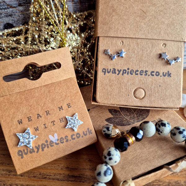 Silver star-shaped stud earrings displayed on kraft paper cards and a box, alongside a beaded bracelet on a wooden surface. The cards and box are printed with the brand name 'quaypieces.