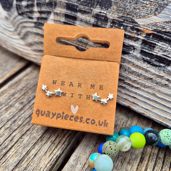 A pair of silver star-shaped stud earrings displayed on a brown cardboard backing card that reads 'WEAR ME WITH quaypieces.co.