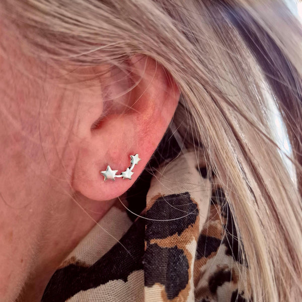 A close-up of a person's ear wearing a silver ear cuff with three star-shaped studs connected by curved bars, along with a leopard-print scarf.