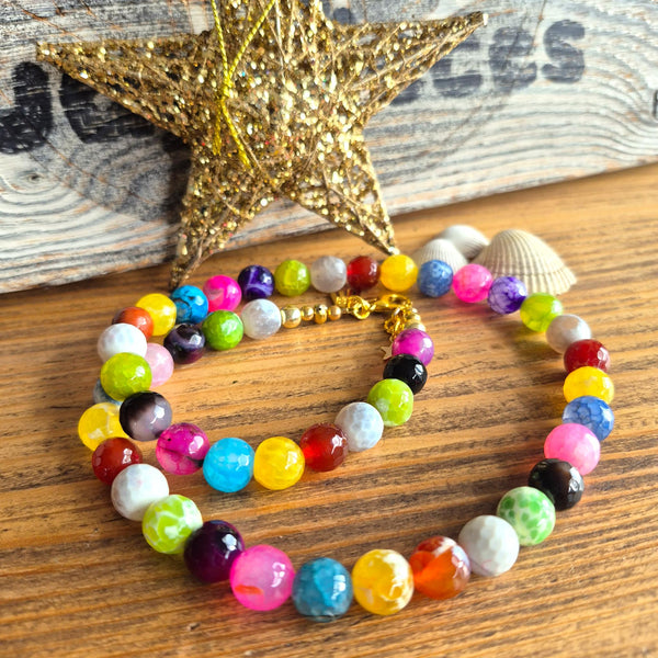 A multicolor beaded necklace and matching bracelet displayed on a wooden surface, styled with a gold glitter star and a small seashell. The beads are in many different bright colors including pink, blue, yellow, green, purple, orange, and white, with some beads showing a marbled pattern.