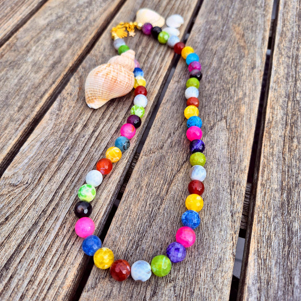 A colorful beaded necklace made of faceted round beads in many bright colors, arranged on weathered wooden planks with two seashells beside it.