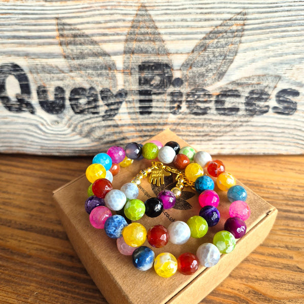 A set of colorful beaded bracelets with a gold elephant charm, displayed on a small brown box in front of a rustic wooden sign that reads 'Quay Pieces'.