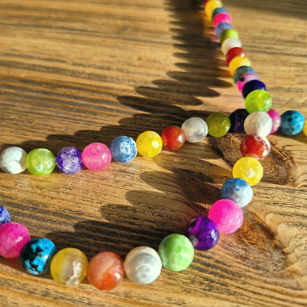 A strand of colorful round gemstone beads arranged in loops on a wooden tabletop, with sunlight casting shadows across the surface.