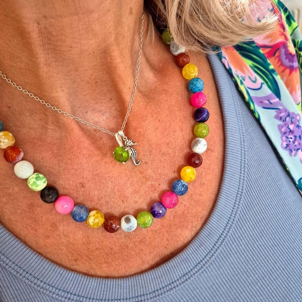 A close-up of a person's neck and chest wearing two necklaces: a silver chain necklace with a small green dragon pendant, and a colorful beaded necklace with round multicolored beads.