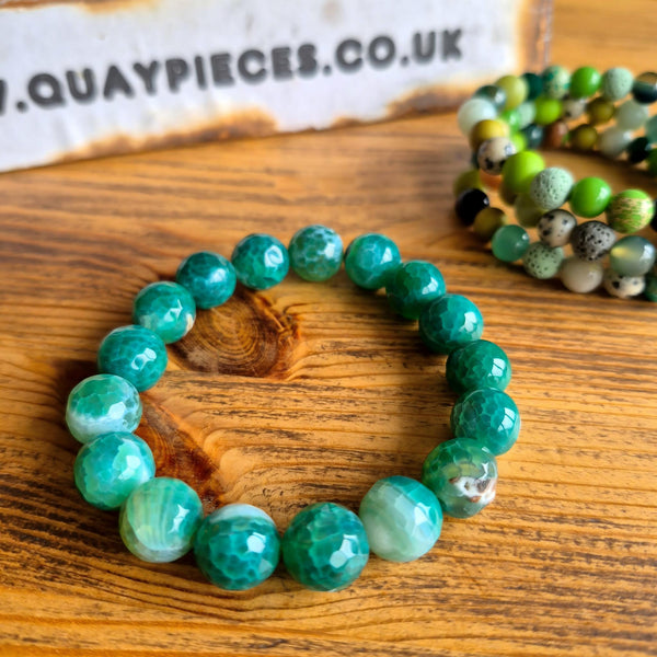 A beaded stretch bracelet made of faceted green marbled beads, displayed on a wooden surface next to a second multicolor beaded bracelet. A branded cardboard box is partially visible in the background.