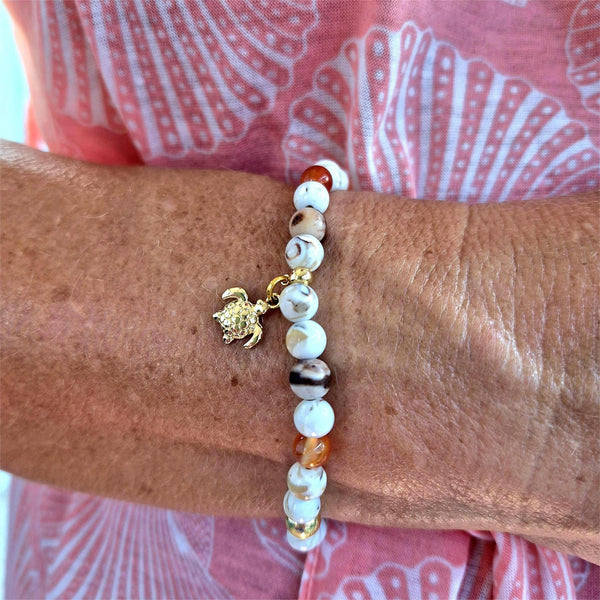 Beaded bracelet with a turtle charm on a wrist, against a pink and white patterned fabric background.