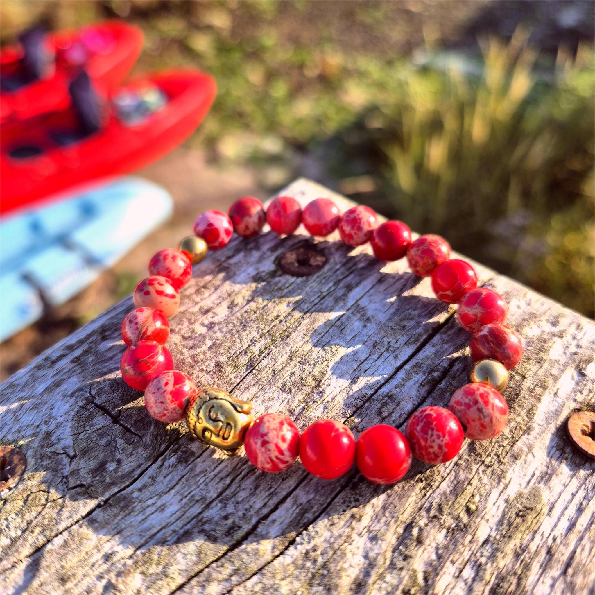 Red beaded bracelet with a gold clasp on a wooden surface, with blurred kayaks in the background.