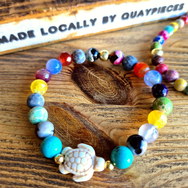 Colorful beaded bracelet with a turtle charm on a wooden surface, featuring the brand 'Quaypieces'.